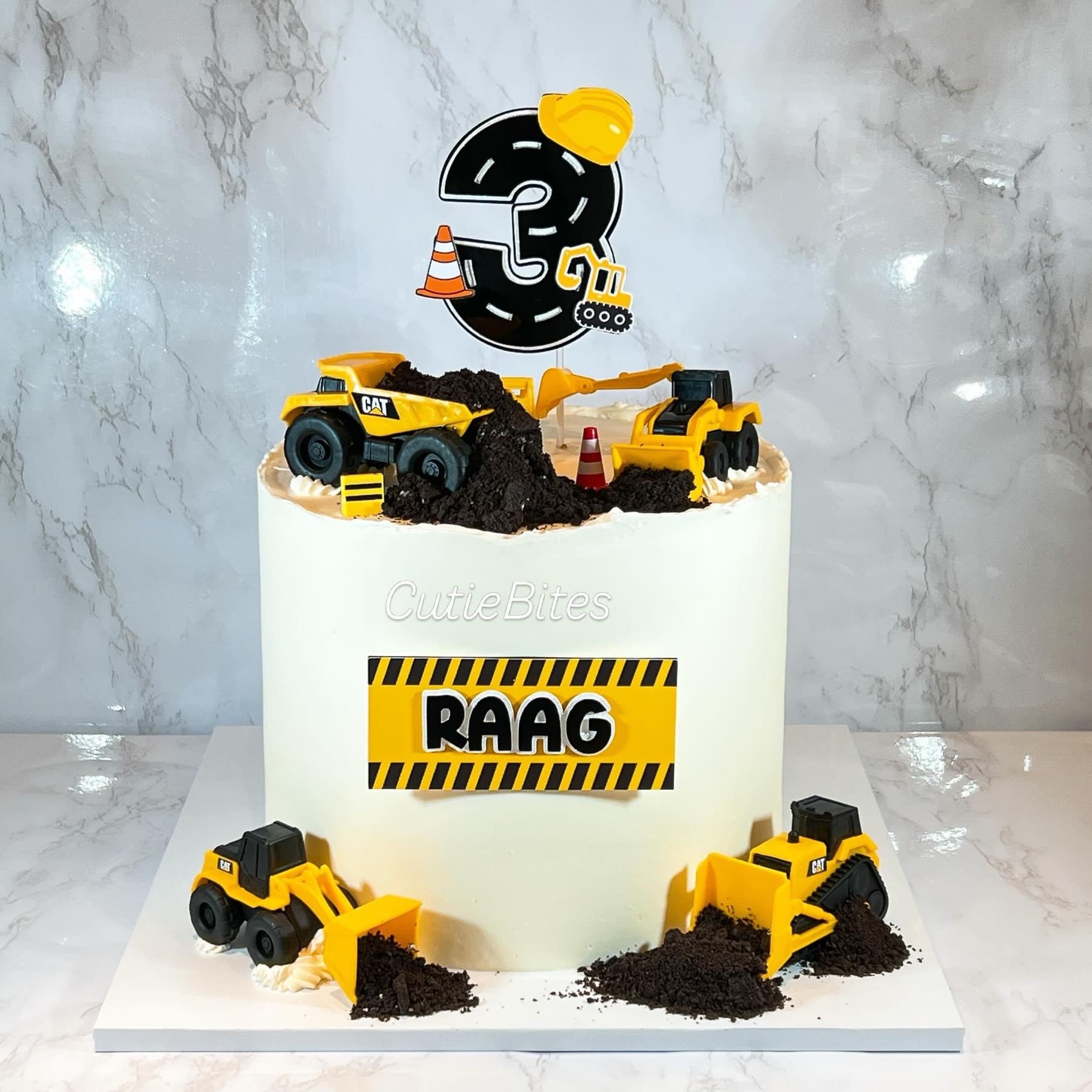 Construction digger birthday cake with yellow trucks, crumb soil and hazard-tape name — Cutie Bites, Orpington